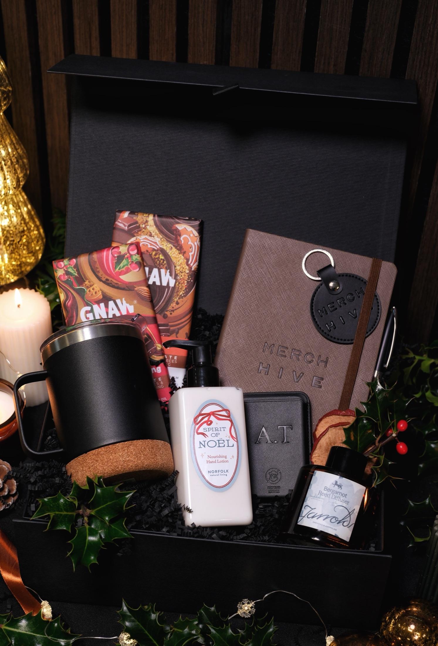 Black Merch Hive gift box with a branded notebook, travel mug, chocolate, hand lotion and a candle, surrounded by holly and fairy lights