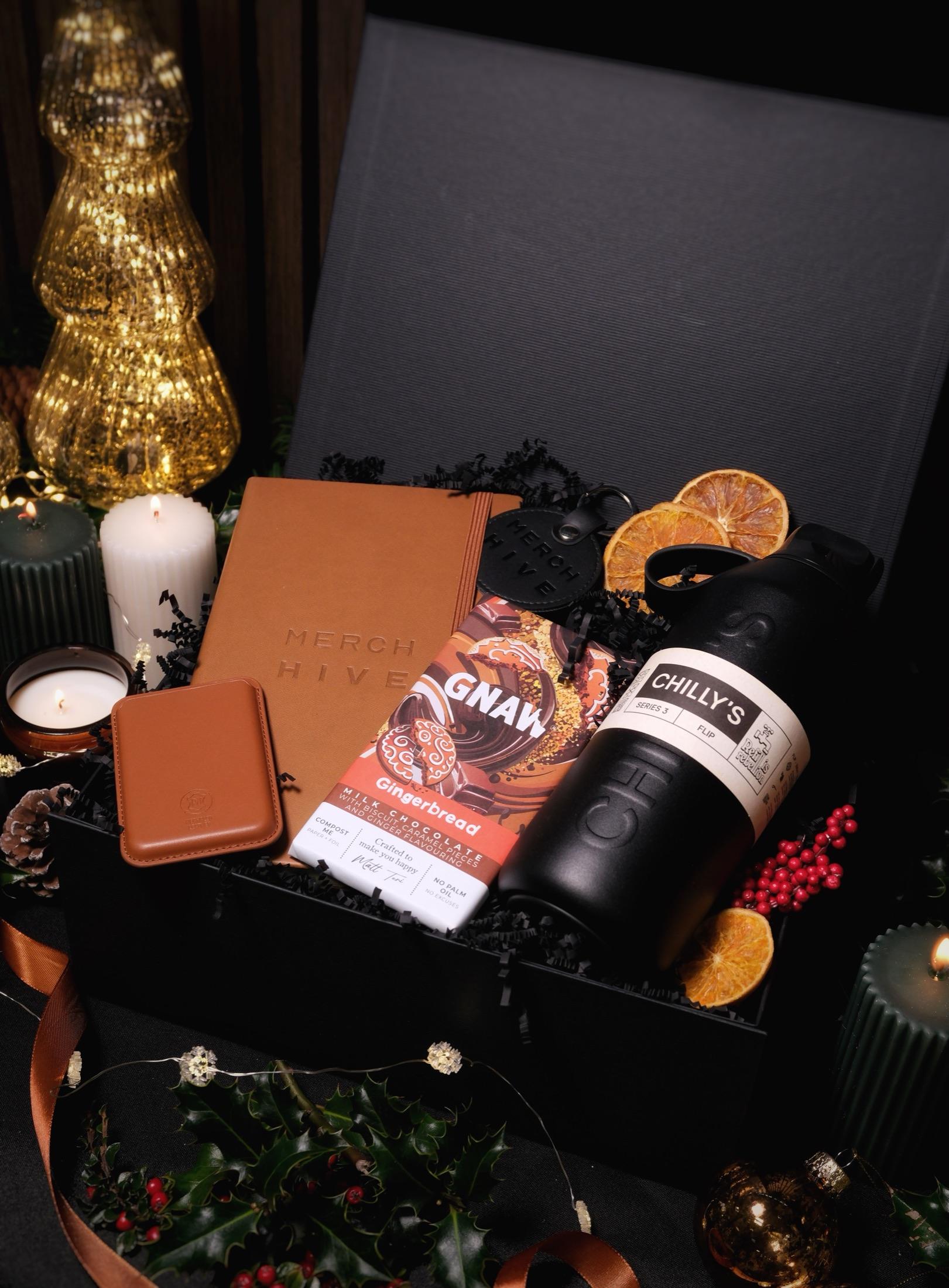 Tan Merch Hive notebook, card holder, gingerbread chocolate and a black water bottle in an open gift box